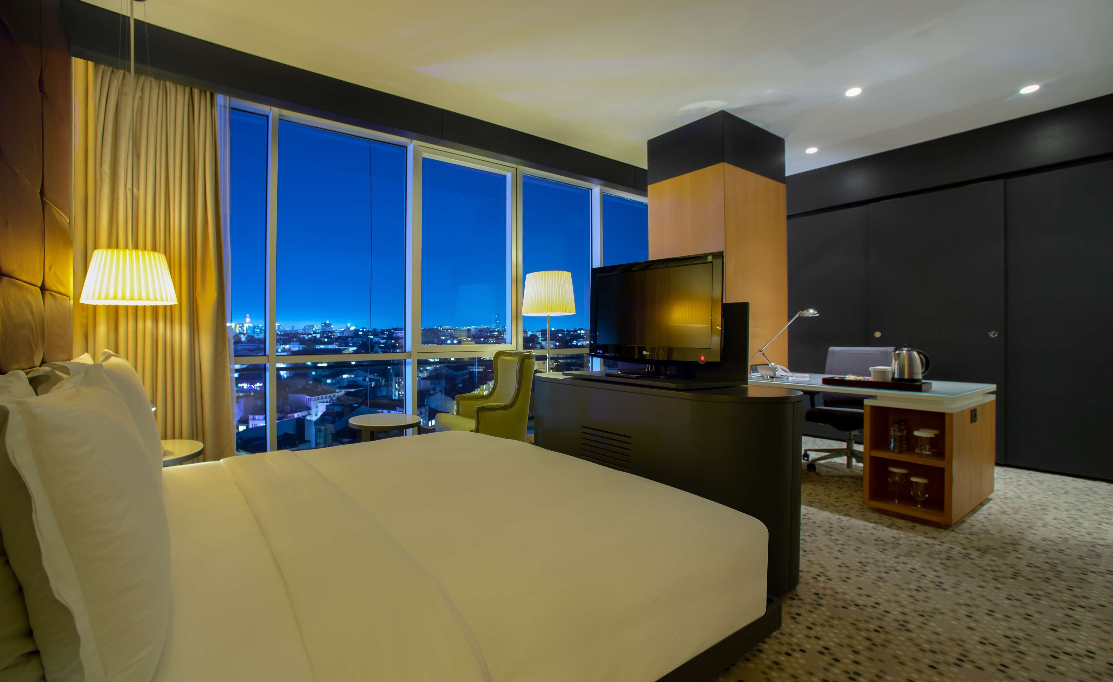 A hotel room with a large bed, a desk with a TV, and a view of the city at night.