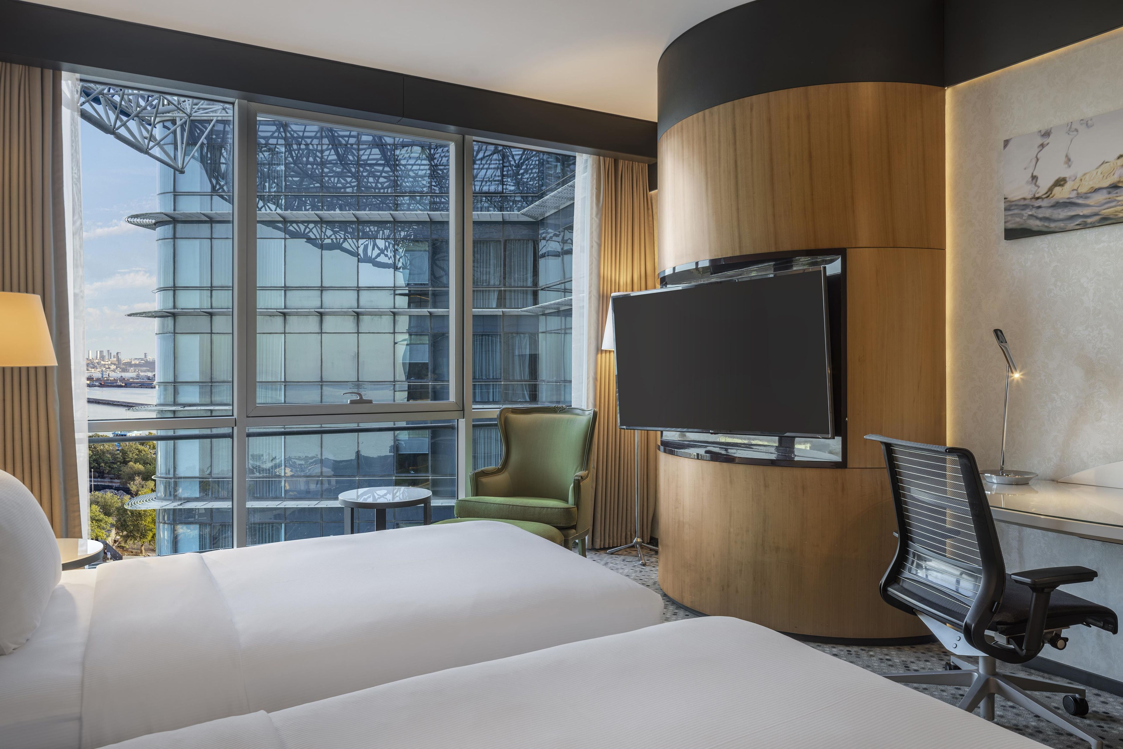 Ein Hotelzimmer mit einem großen Bett, einem an einer Holzvertäfelung montierten Fernseher, einem grünen Sessel, einem kleinen runden Tisch und Blick auf eine Stadtlandschaft durch große Fenster.
