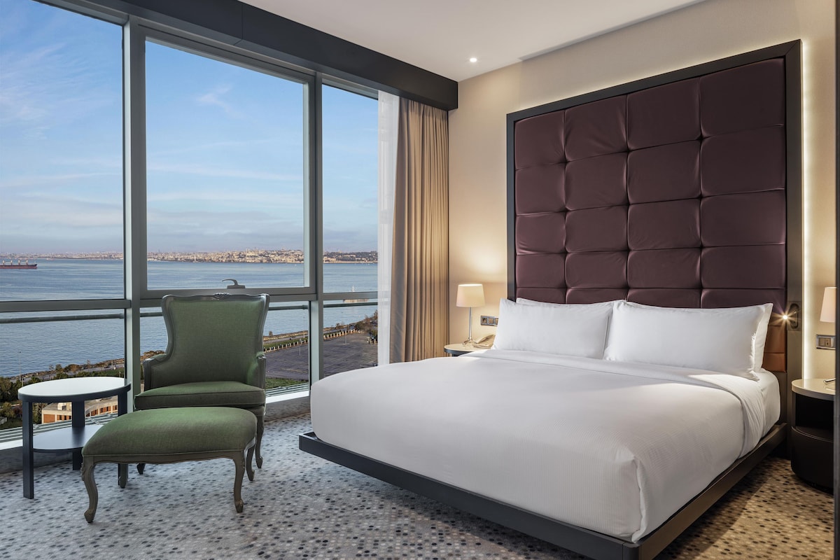 Habitación de hotel con una cama grande, un sillón verde, una mesita y vistas a un cuerpo de agua.