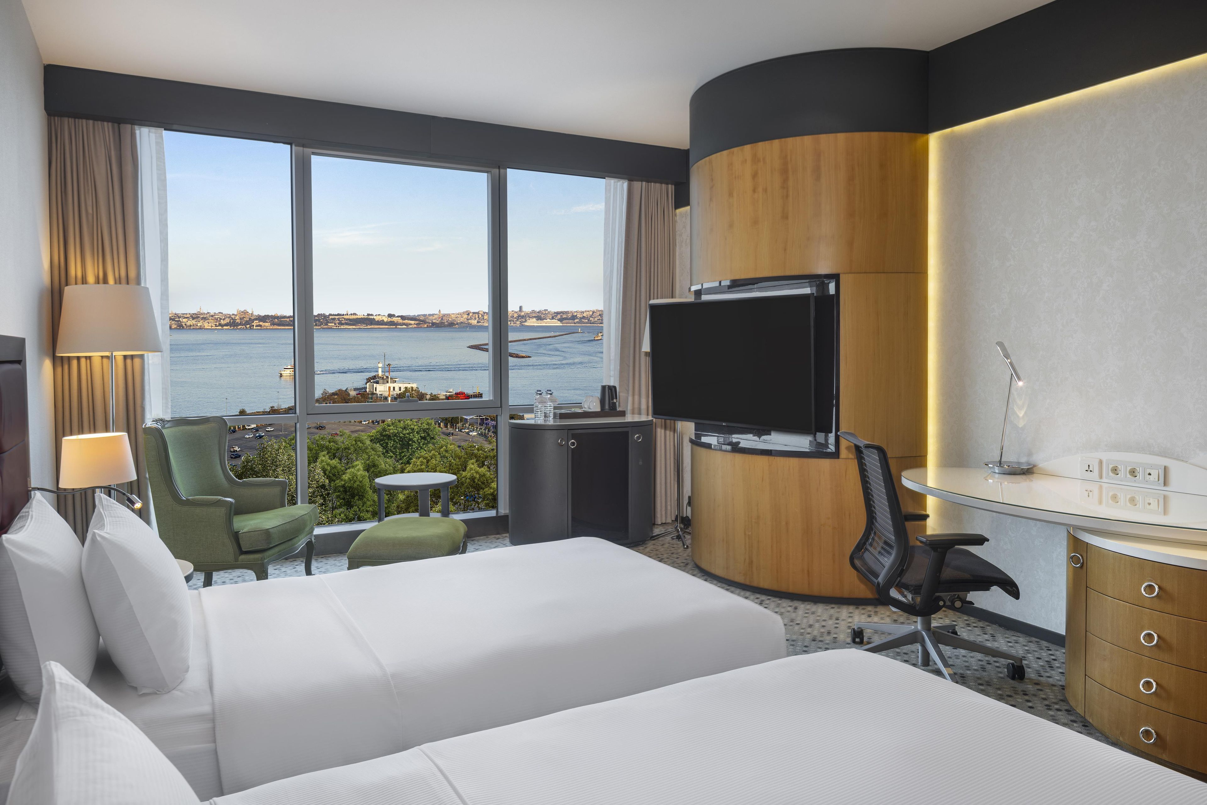 A hotel room with two beds, a desk, a chair, a TV, and a view of the sea.