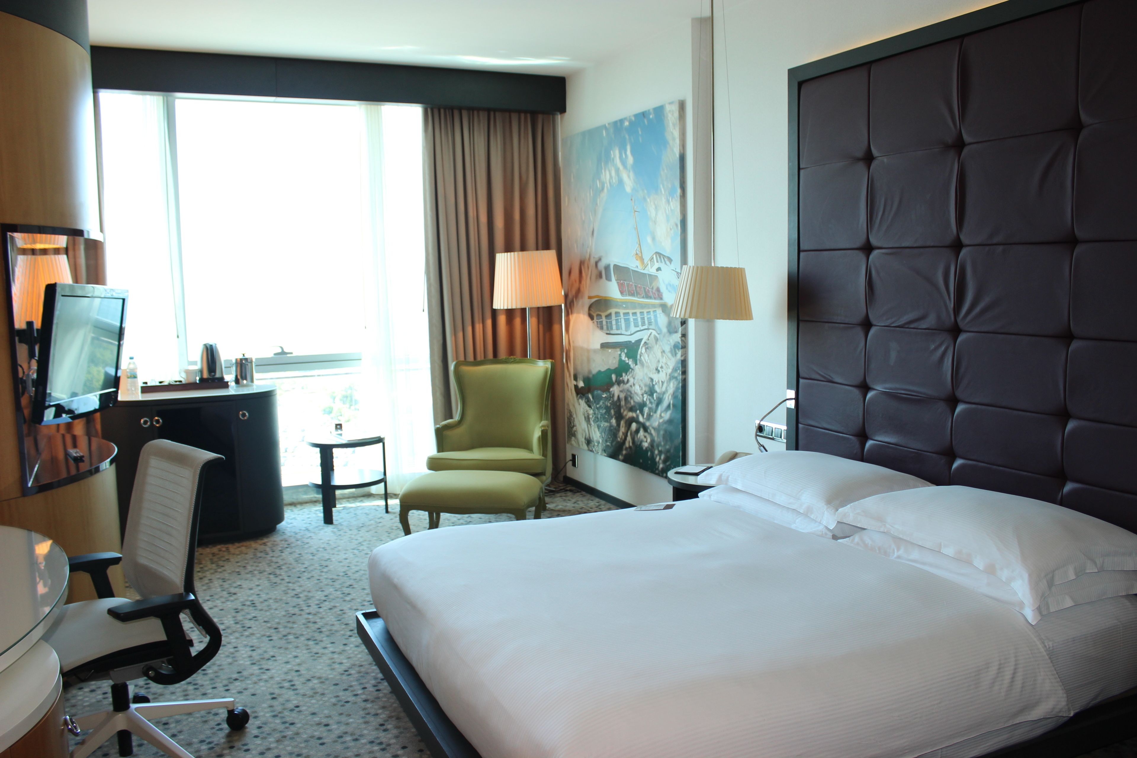 A hotel room with a large bed, a desk with a computer, a green armchair, and a painting on the wall.