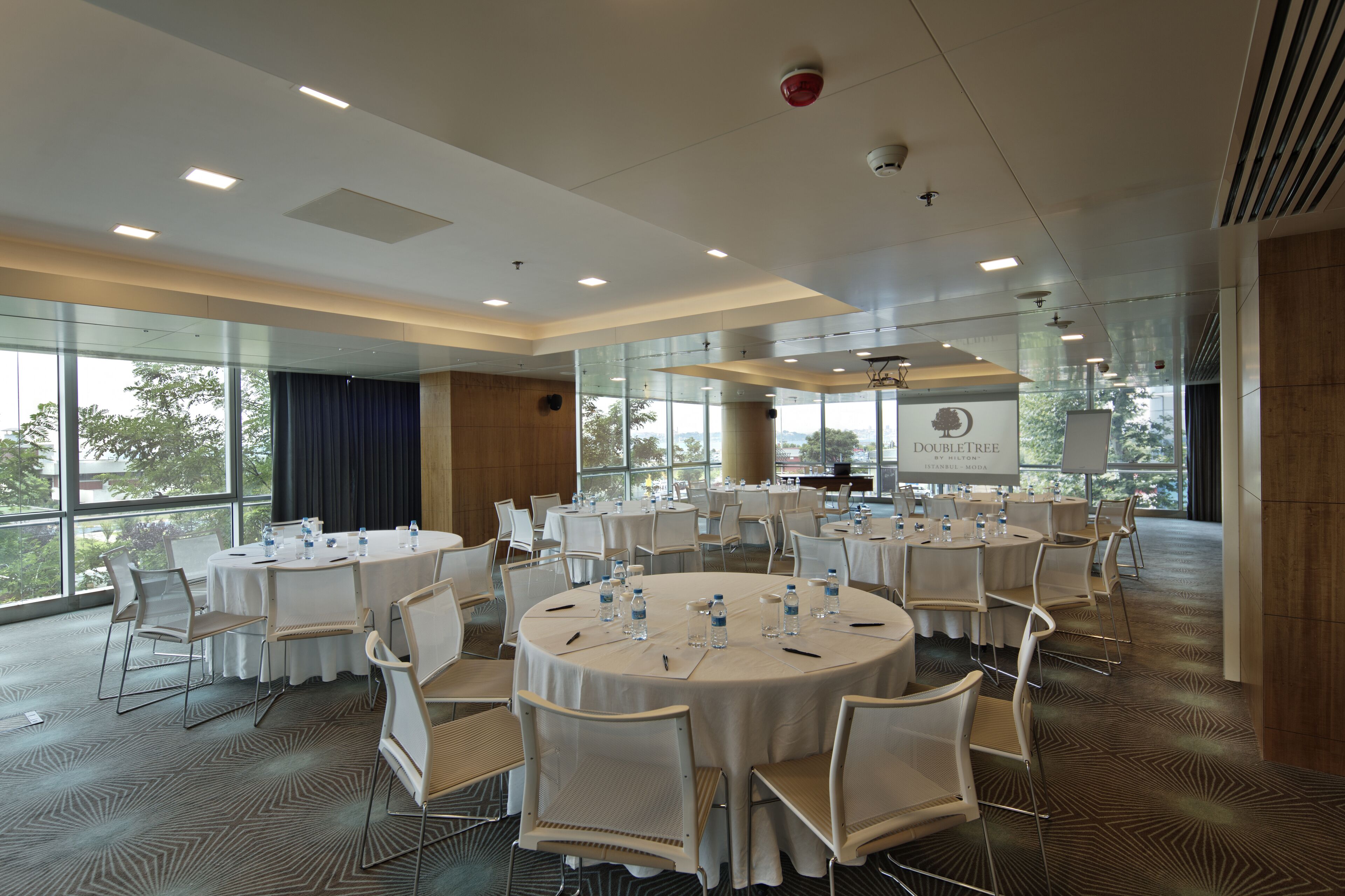 Un salón de conferencias con mesas redondas, sillas y botellas de agua, ventanas amplias y un letrero con la leyenda 'DOUBLE TREE'.