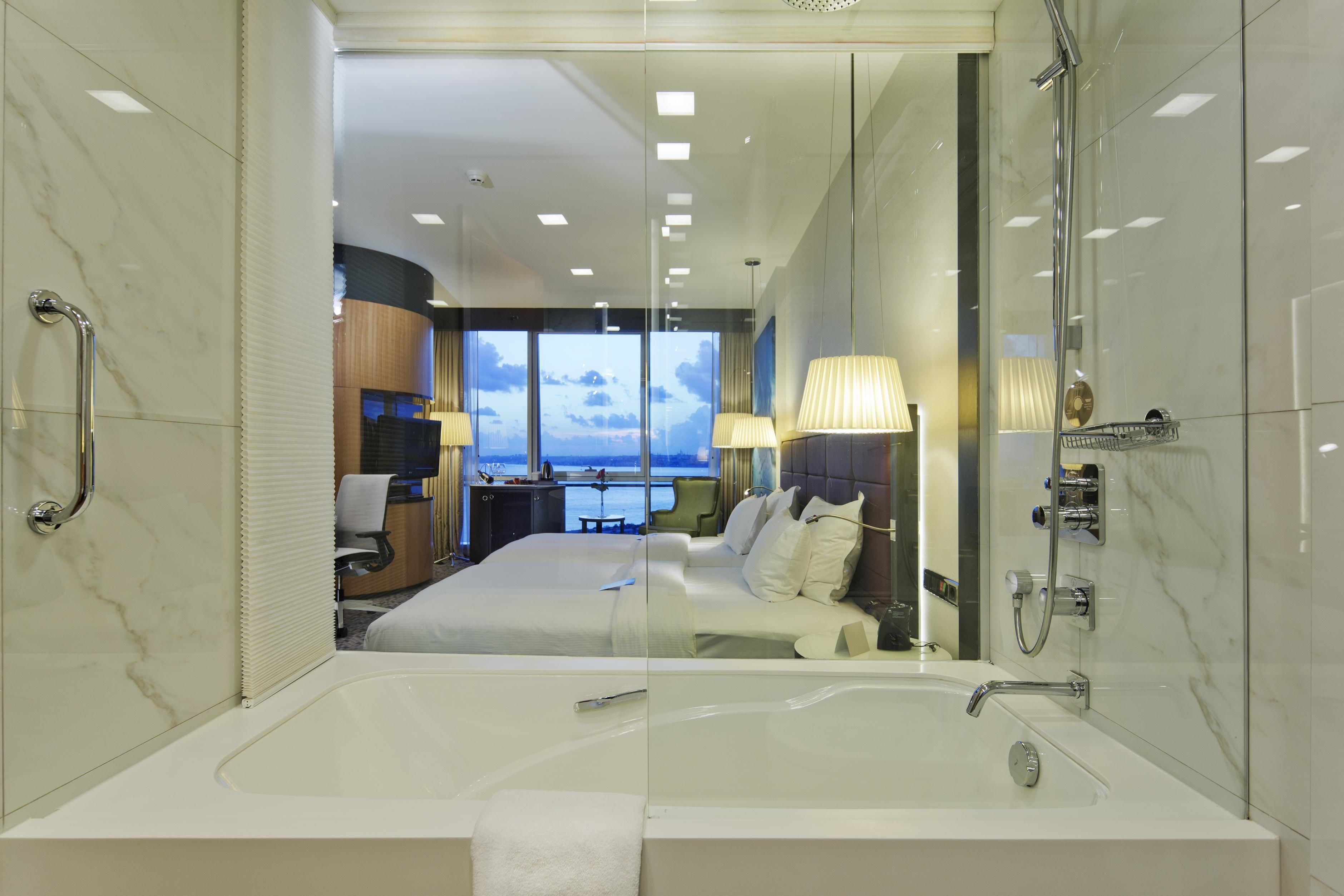 A modern hotel room with a large bed, a bathtub, and a view of the sea through the window.
