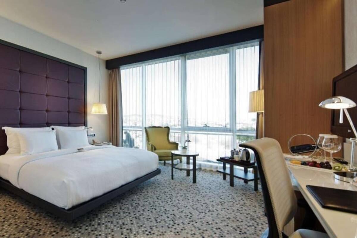 A hotel room with a large bed, two bedside tables, a desk with a chair, a TV, and a large window with curtains.