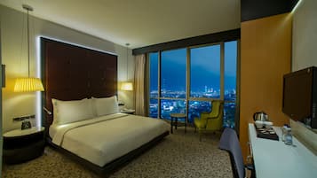 A hotel room with a large bed, a desk, a chair, a TV, and a view of the city at night.