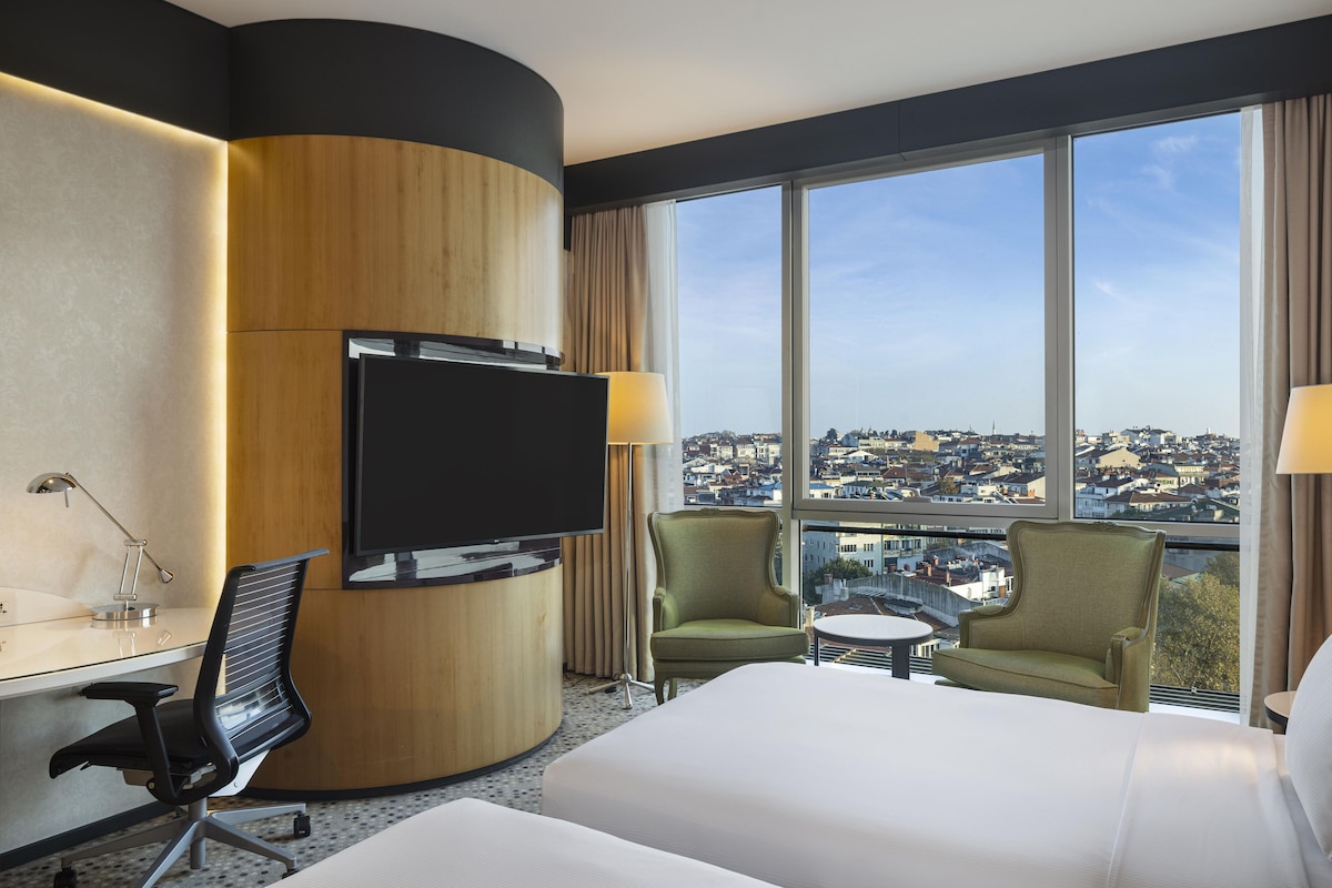 Habitación de hotel con cama, escritorio con silla, televisión y vista a un paisaje urbano a través de amplias ventanas.