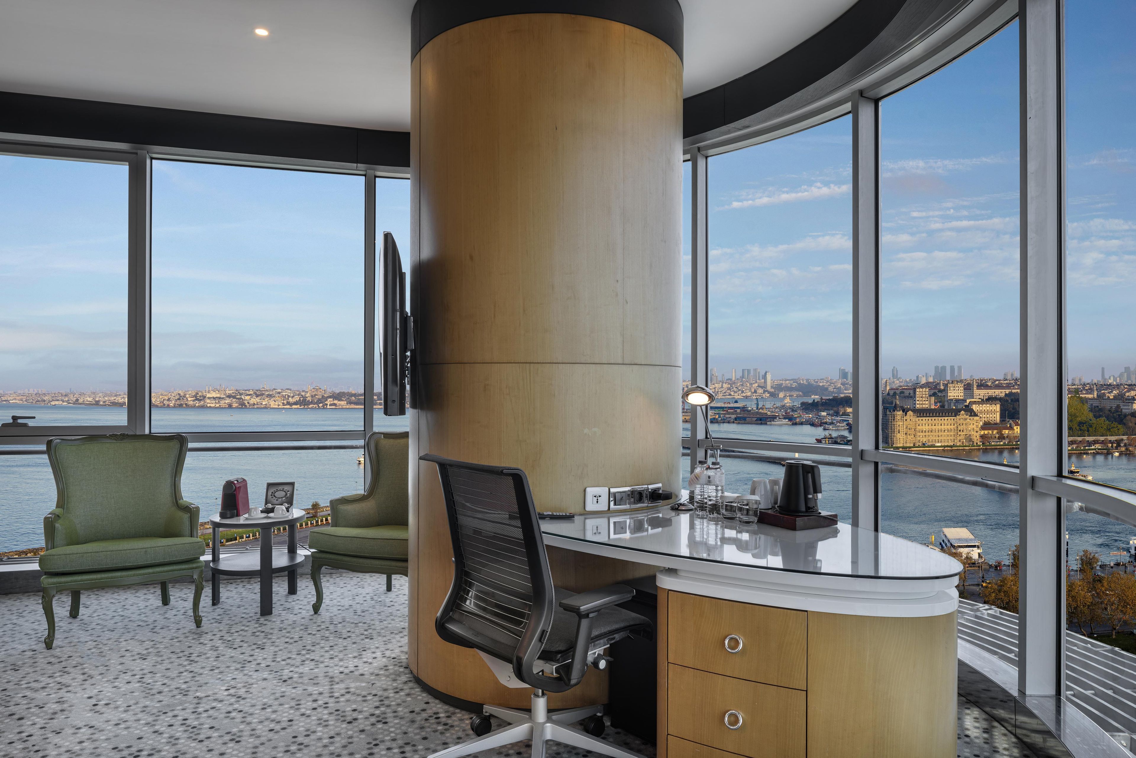 Un espacio de oficina moderno con un escritorio grande, dos sillas, un ventanal con vistas a la ciudad y una columna alta.