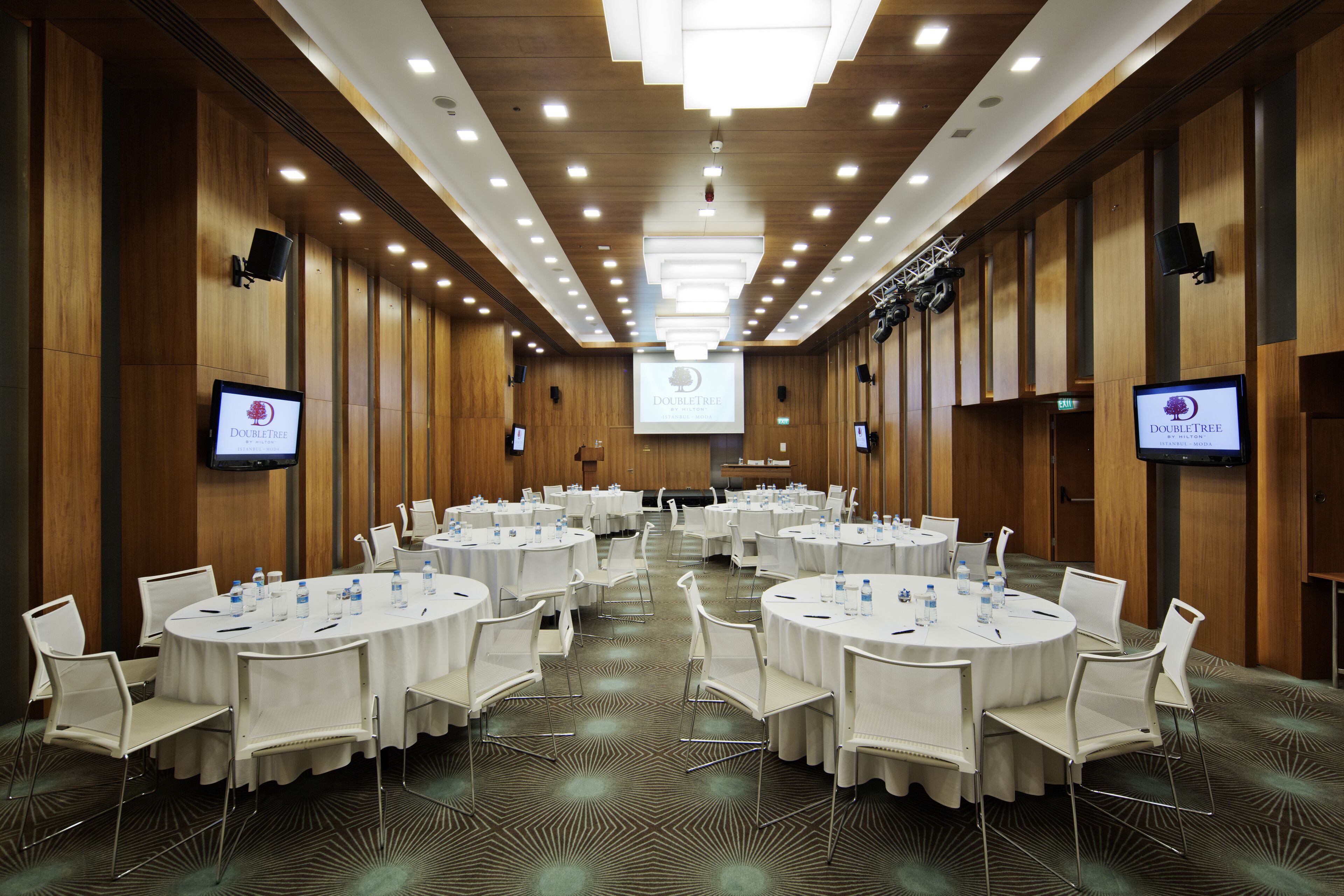 A conference room with round tables, white chairs, and a large screen at the front.
