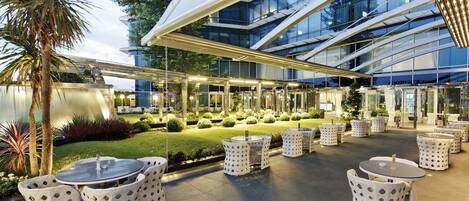 An outdoor patio with round tables and white chairs, surrounded by a well-maintained garden and a modern building with glass windows.