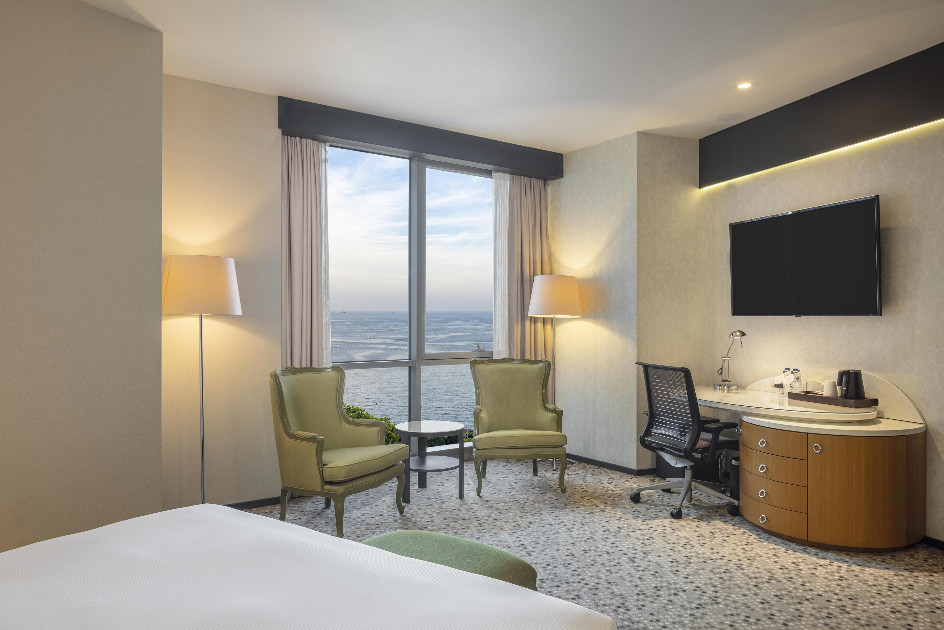 Una camera d'albergo con un letto, due sedie, una scrivania con un computer, una TV a schermo piatto e vista sul mare.