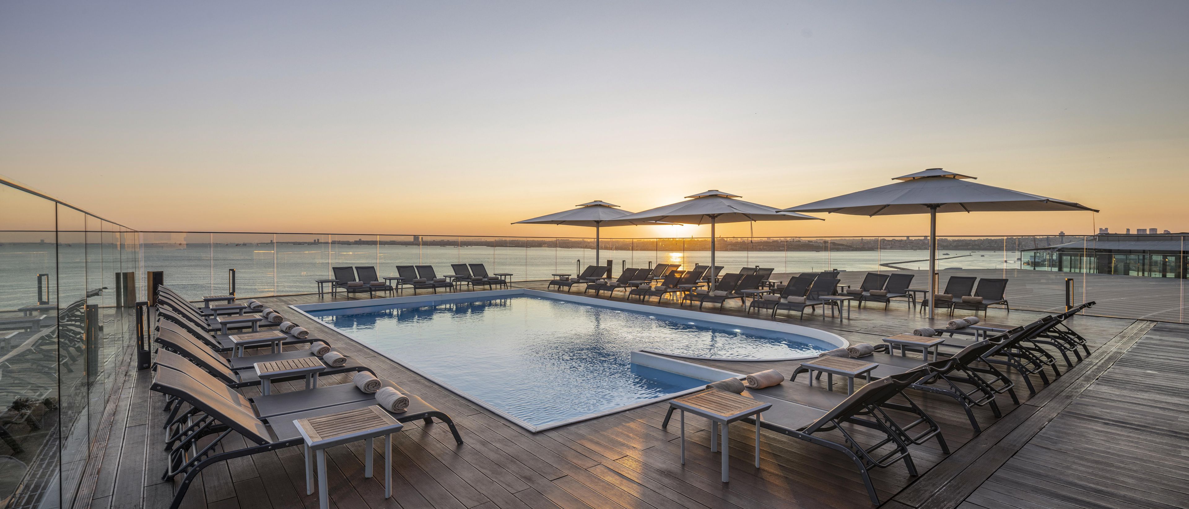 A rooftop pool area with lounge chairs and umbrellas, overlooking a body of water.
