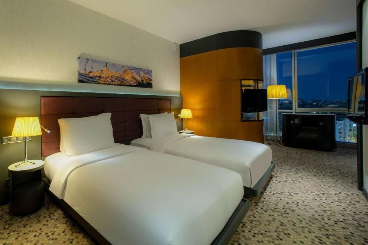A hotel room with two beds, a large window with a city view, a painting on the wall, and a TV on a stand.
