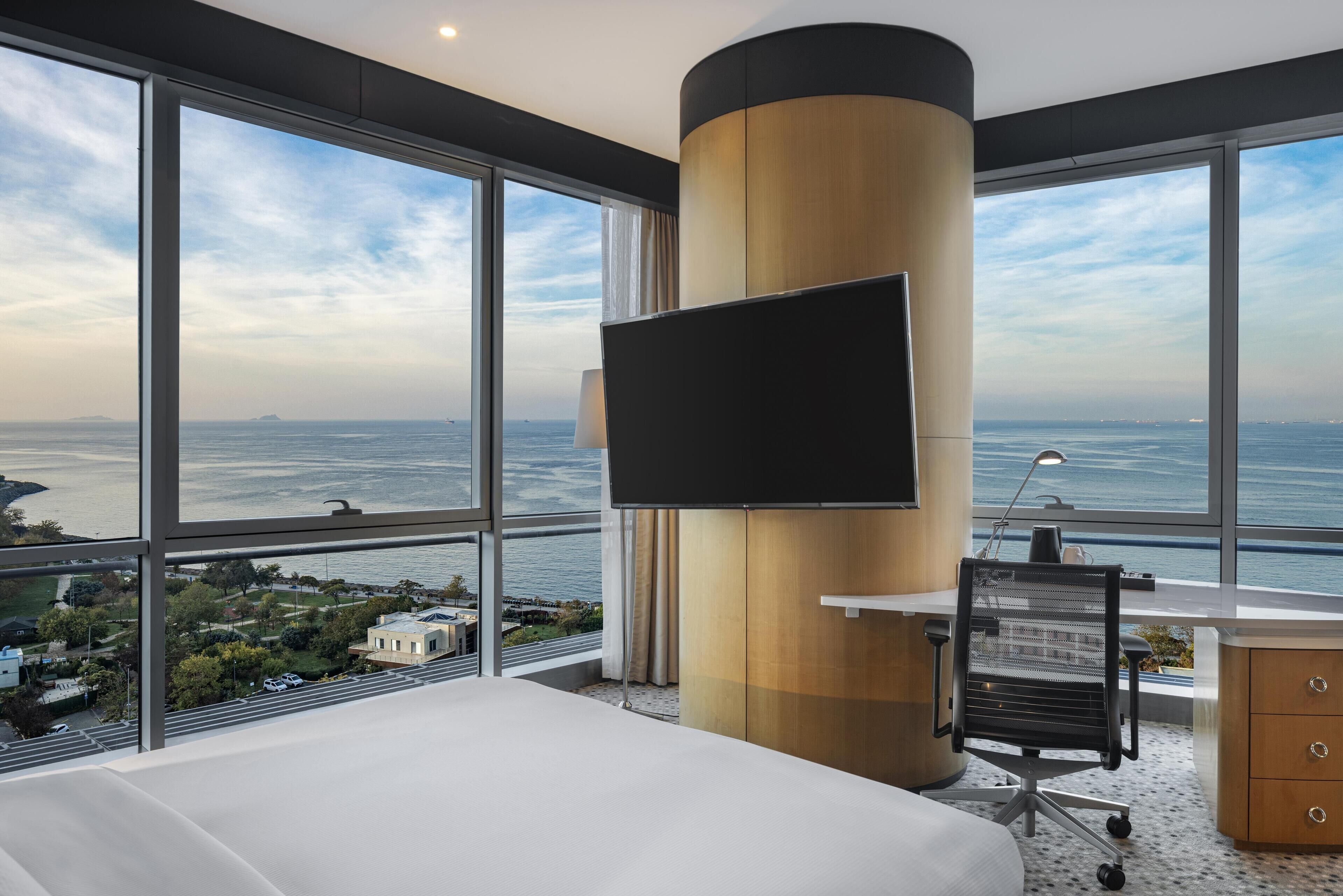 Habitación de hotel con una cama grande, un escritorio con una silla, un televisor fijado a un pilar de madera y vistas al mar y al paisaje urbano.