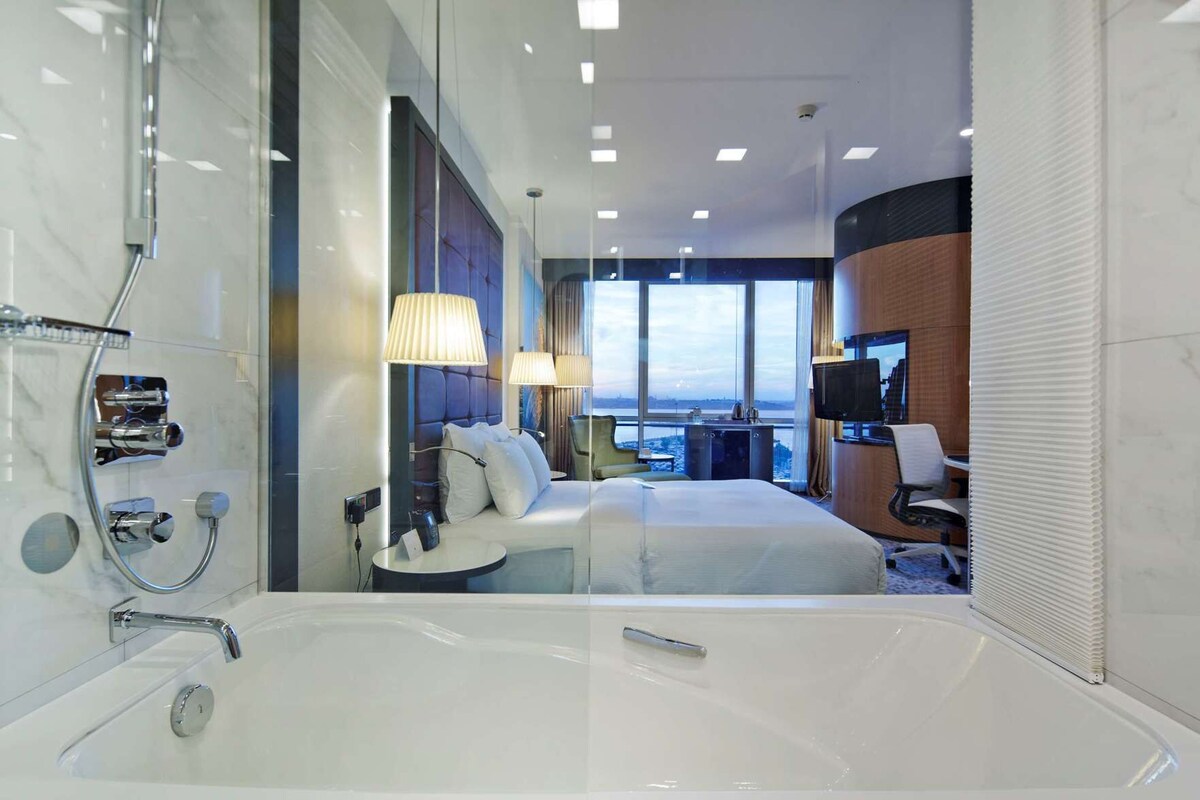 Une chambre d’hôtel moderne dotée d’une grande baignoire, d’un lit avec du linge de maison blanc, d’un bureau avec un ordinateur et d’une grande fenêtre offrant une vue sur la ville.