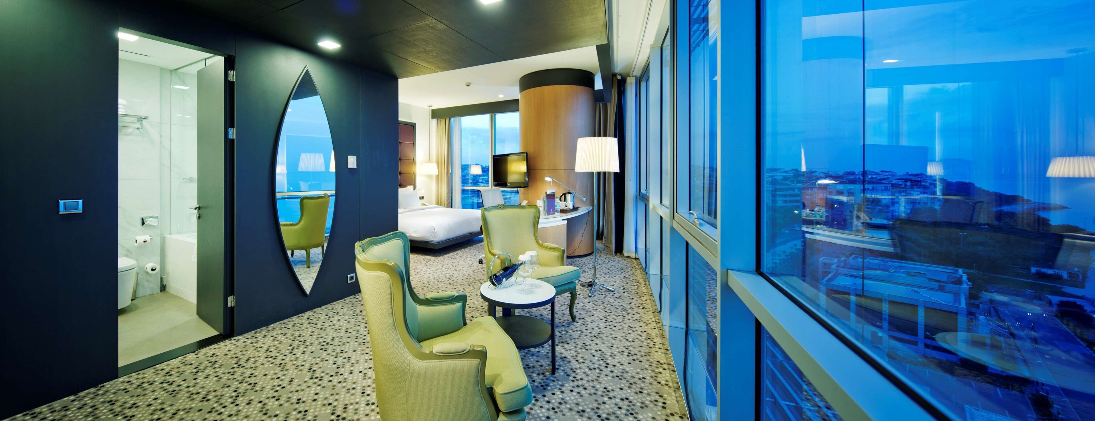 A hotel room with a large window, a bed, a desk, and a seating area with chairs and a table.