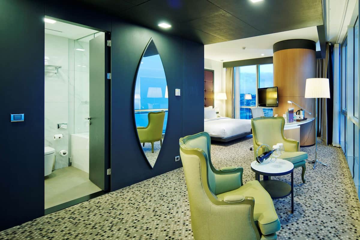 A hotel room with a large window, a bed, a desk, and a seating area with chairs and a table.