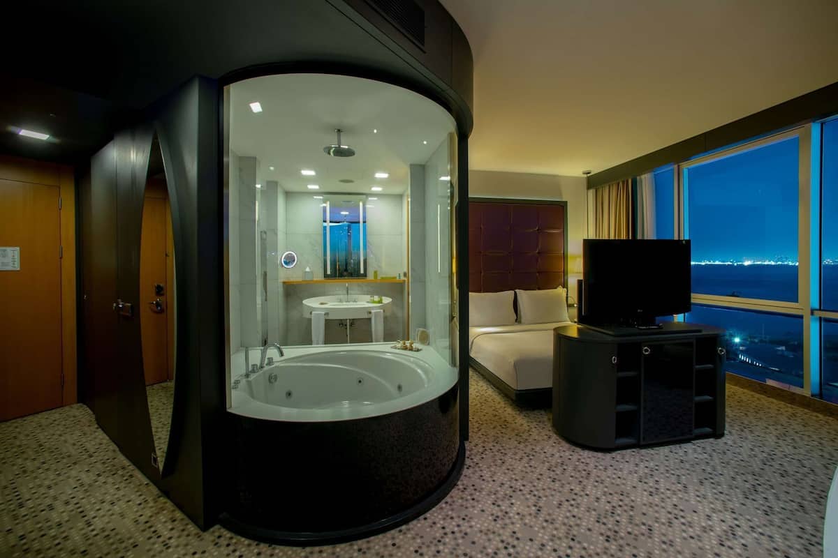 Una habitación con bañera de hidromasaje, televisión, cama y un amplio ventanal con vistas a la ciudad.