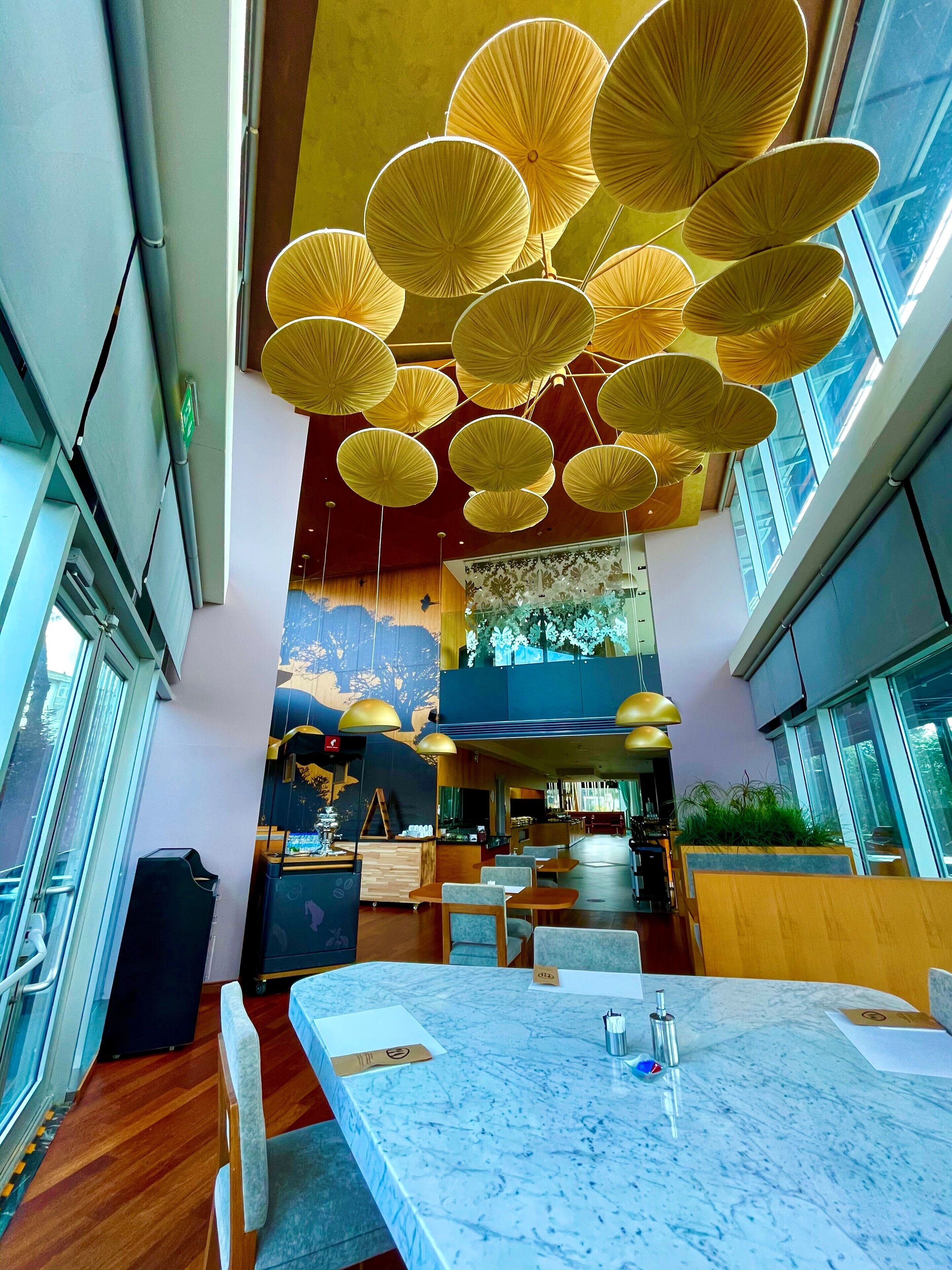 Un restaurant moderne avec un plafond haut, des lampes jaunes suspendues et une vue sur le paysage urbain grâce à de grandes fenêtres.