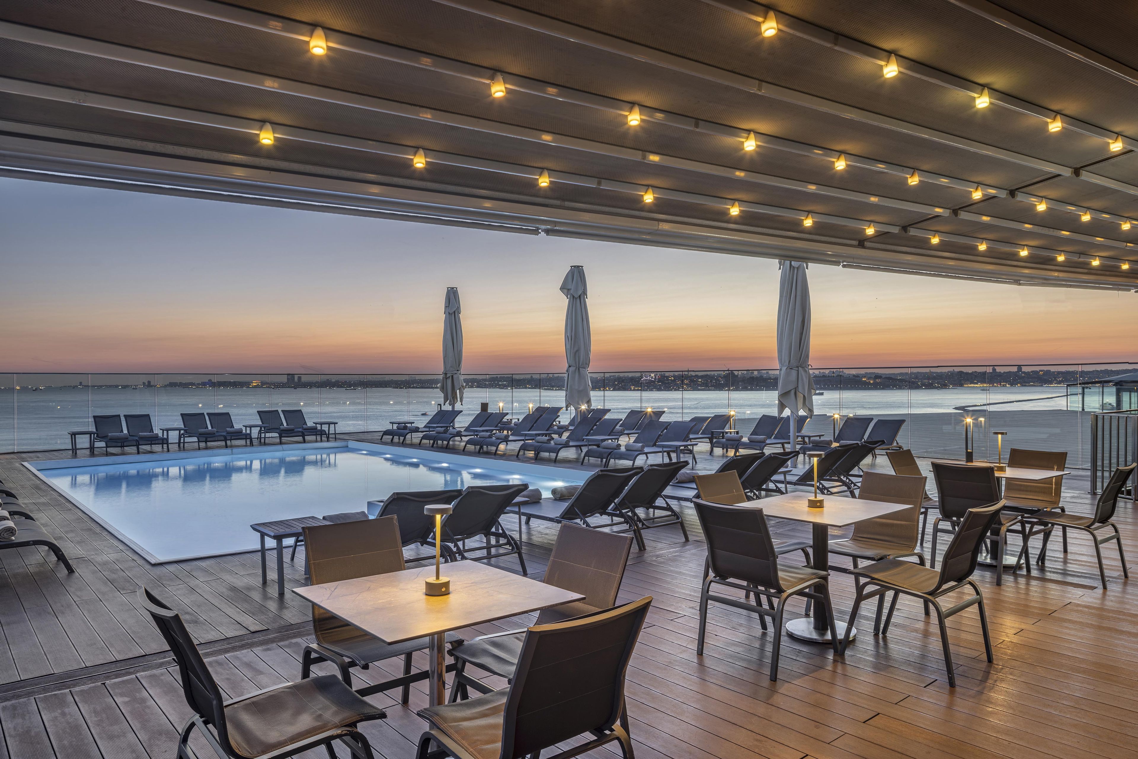 A rooftop pool area with lounge chairs and tables, overlooking a body of water during sunset.