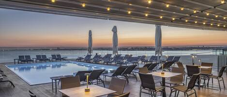 A rooftop pool area with lounge chairs and tables, overlooking a body of water during sunset.