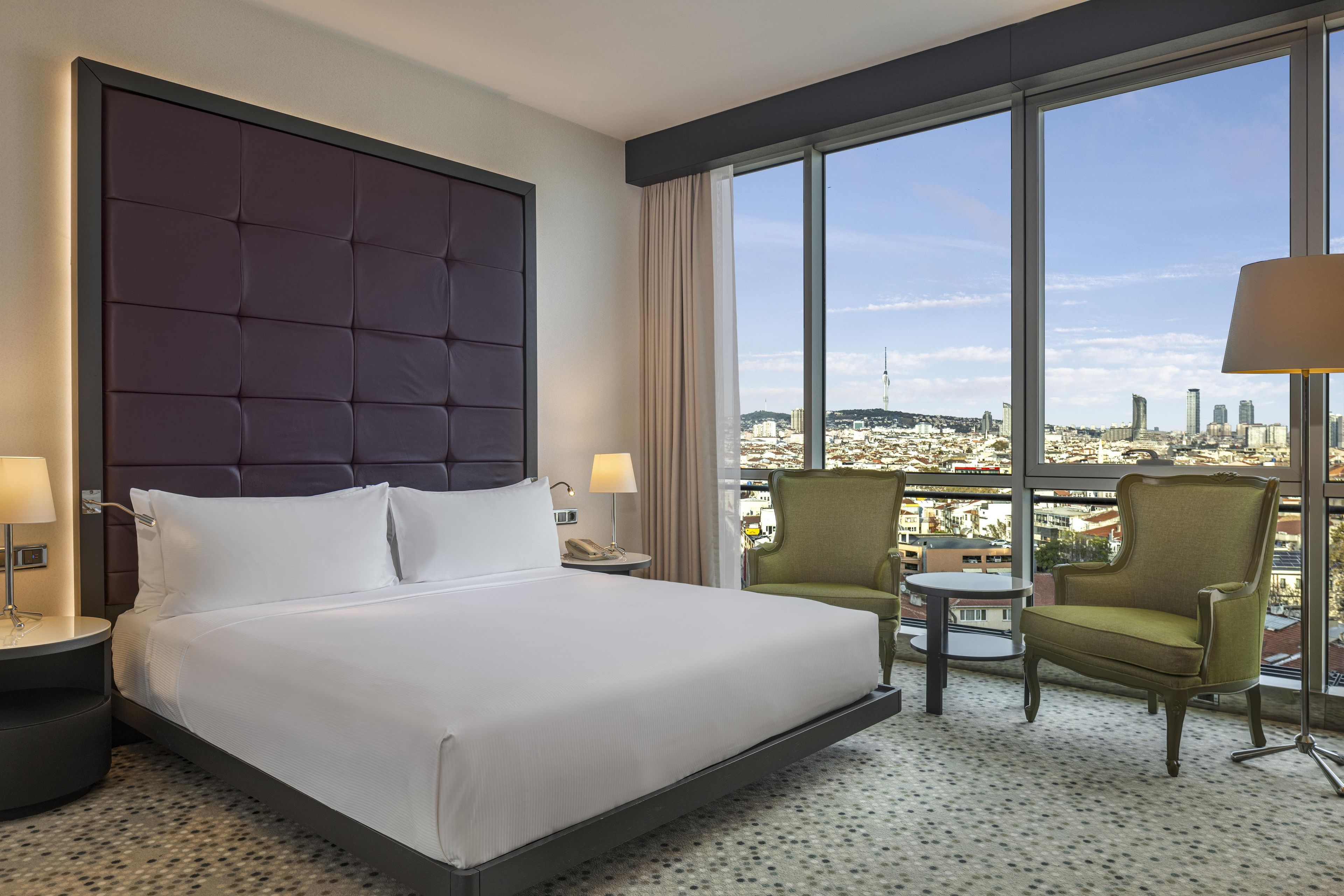 A hotel room with a large bed, two chairs, a small table, and a view of a cityscape through large windows.