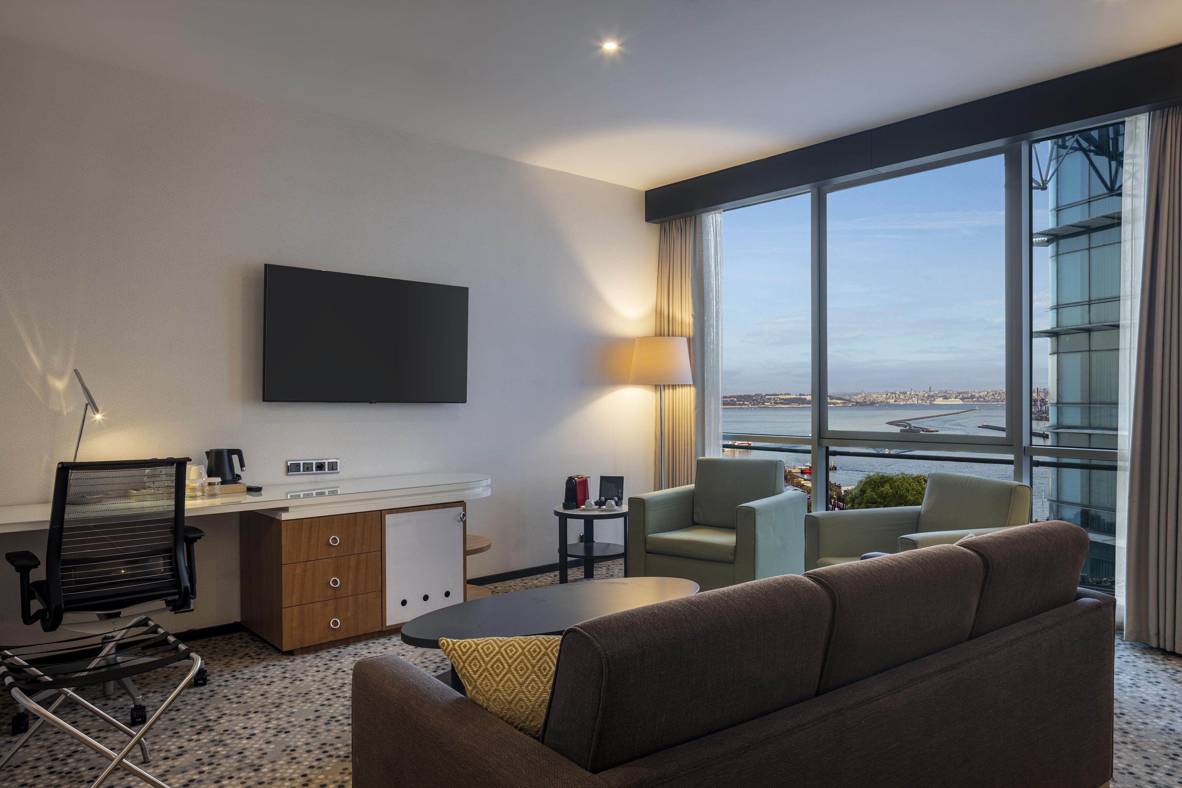 A modern hotel room with a large window, a flat-screen TV, a sofa, a coffee table, and a desk with a lamp.