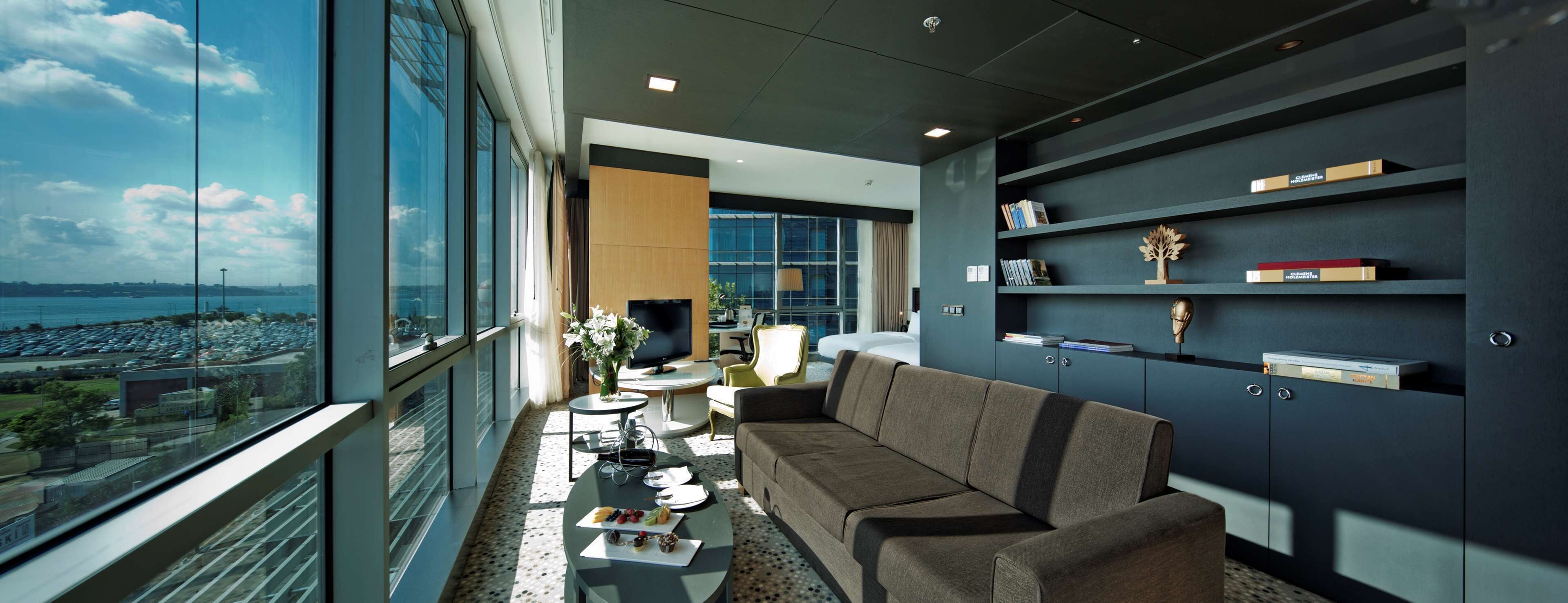 A modern living room with a sofa, a coffee table, and a TV. There are bookshelves with books and decorative items. A large window offers a view of the city and water.