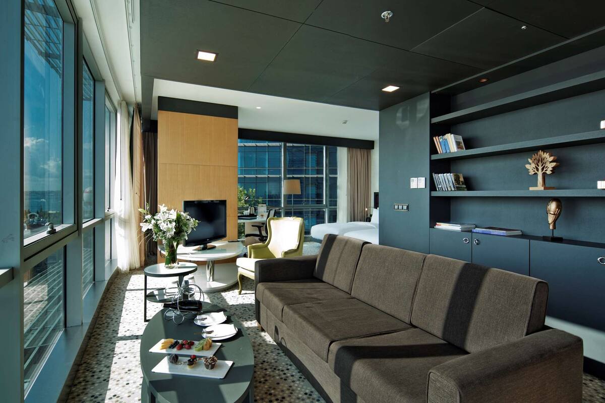 A modern living room with a sofa, a coffee table, and a TV. There are bookshelves with books and decorative items. A large window offers a view of the city and water.