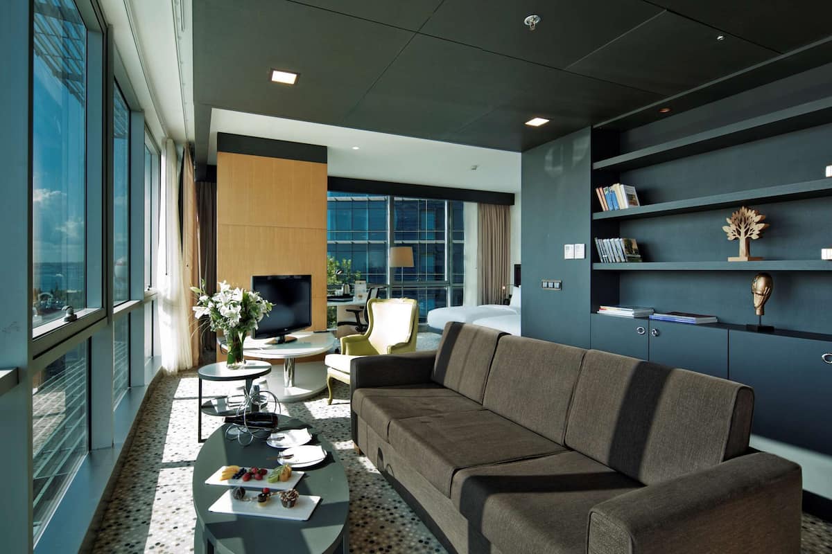 A modern living room with a sofa, a coffee table, and a TV. There are bookshelves with books and decorative items. A large window offers a view of the city and water.