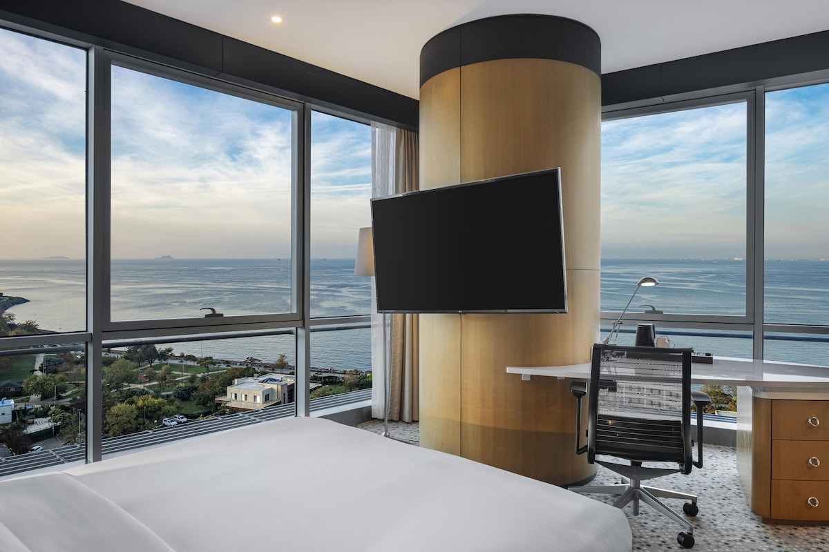 Habitación de hotel con una cama grande, un escritorio con una silla, un televisor fijado a un pilar de madera y vistas al mar y al paisaje urbano.