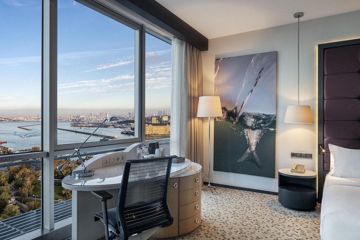 Habitación de hotel con un ventanal grande, un escritorio con una silla, una cama, un cuadro y una mesita de noche con una lámpara.