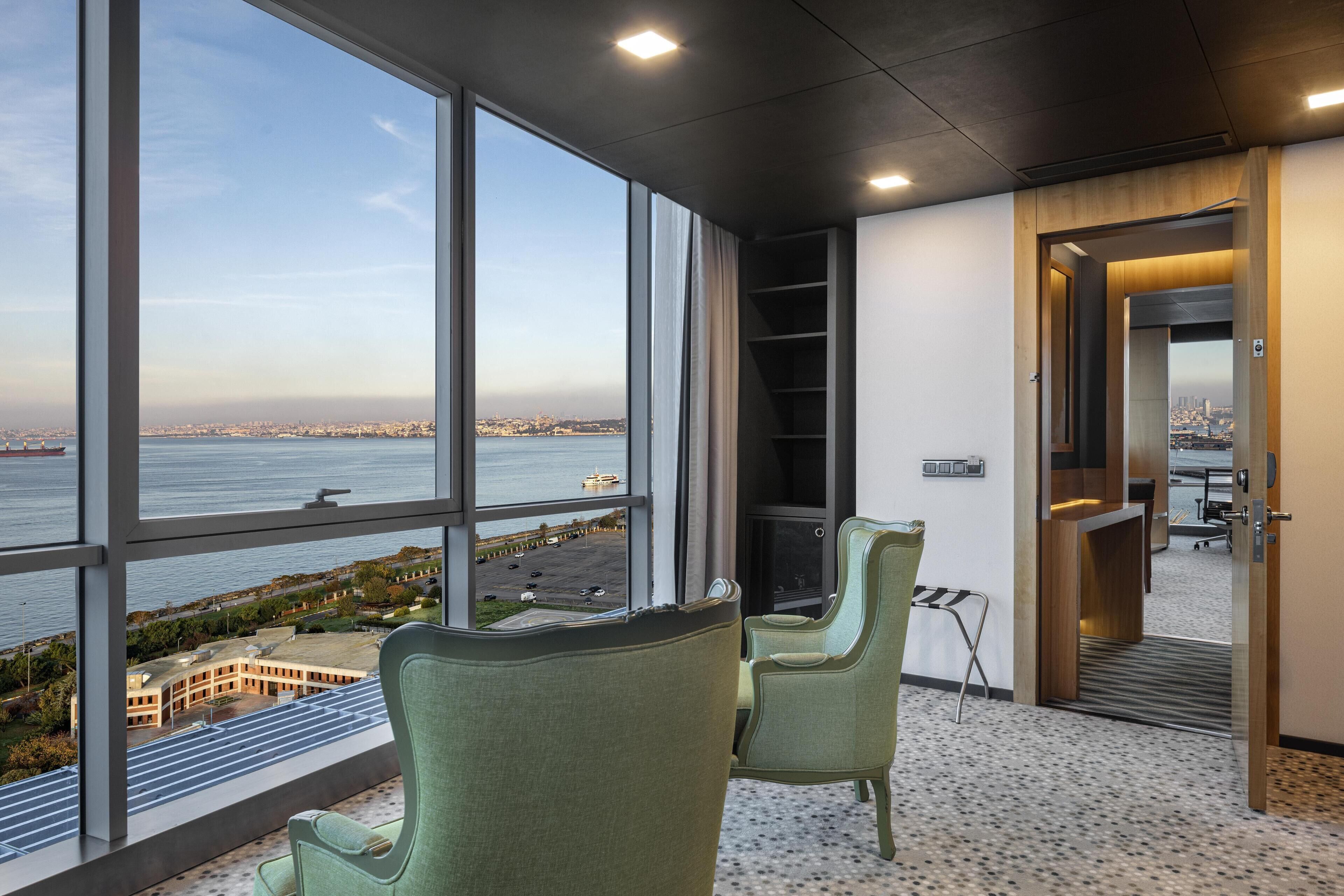 Espacio de oficina moderno con grandes ventanales que dan a una vista panorámica de la ciudad y el agua. Hay dos sillas verdes y un estante empotrado.