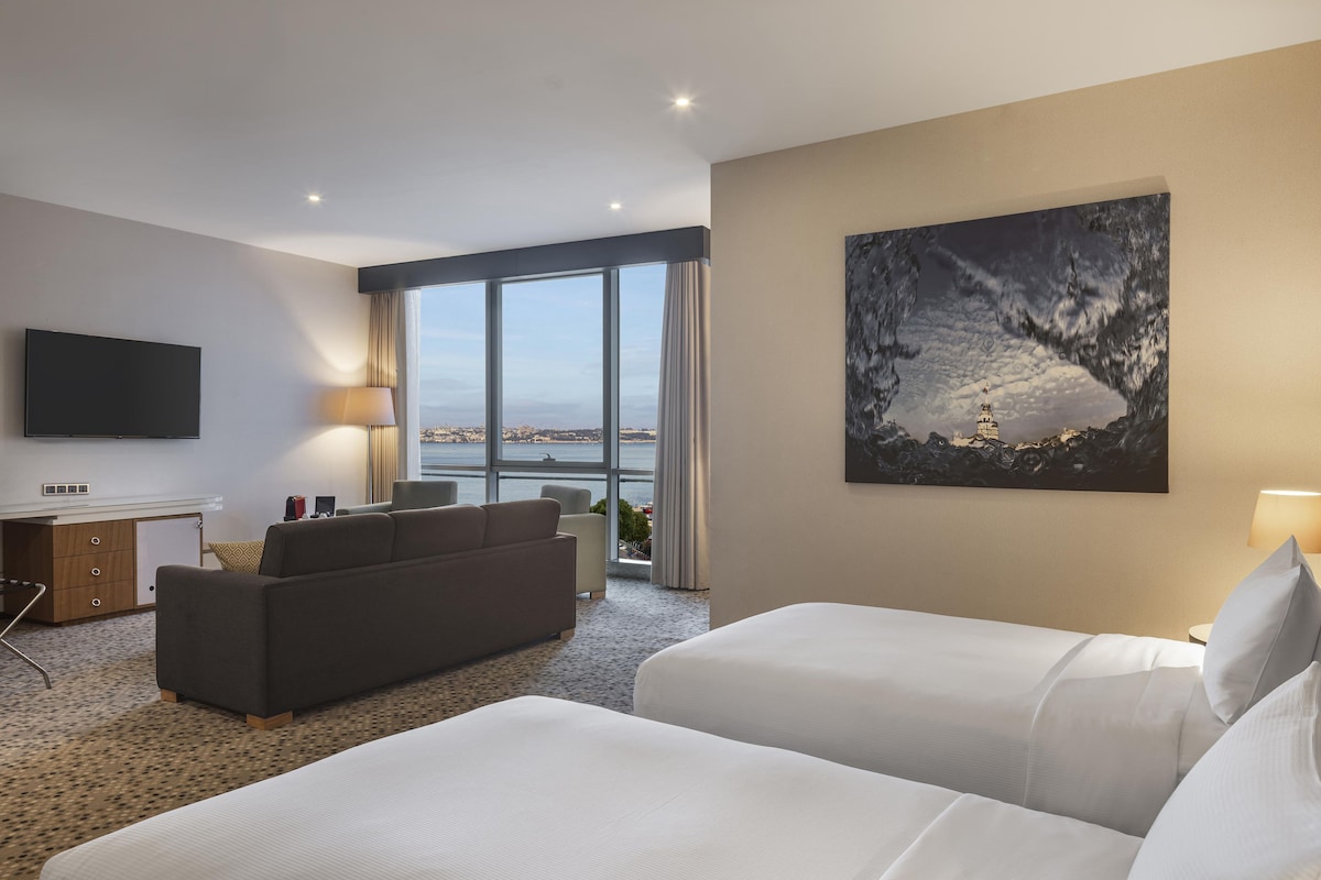 Habitación de hotel con un ventanal grande, televisor de pantalla plana, sofá, dos camas y un cuadro en la pared.
