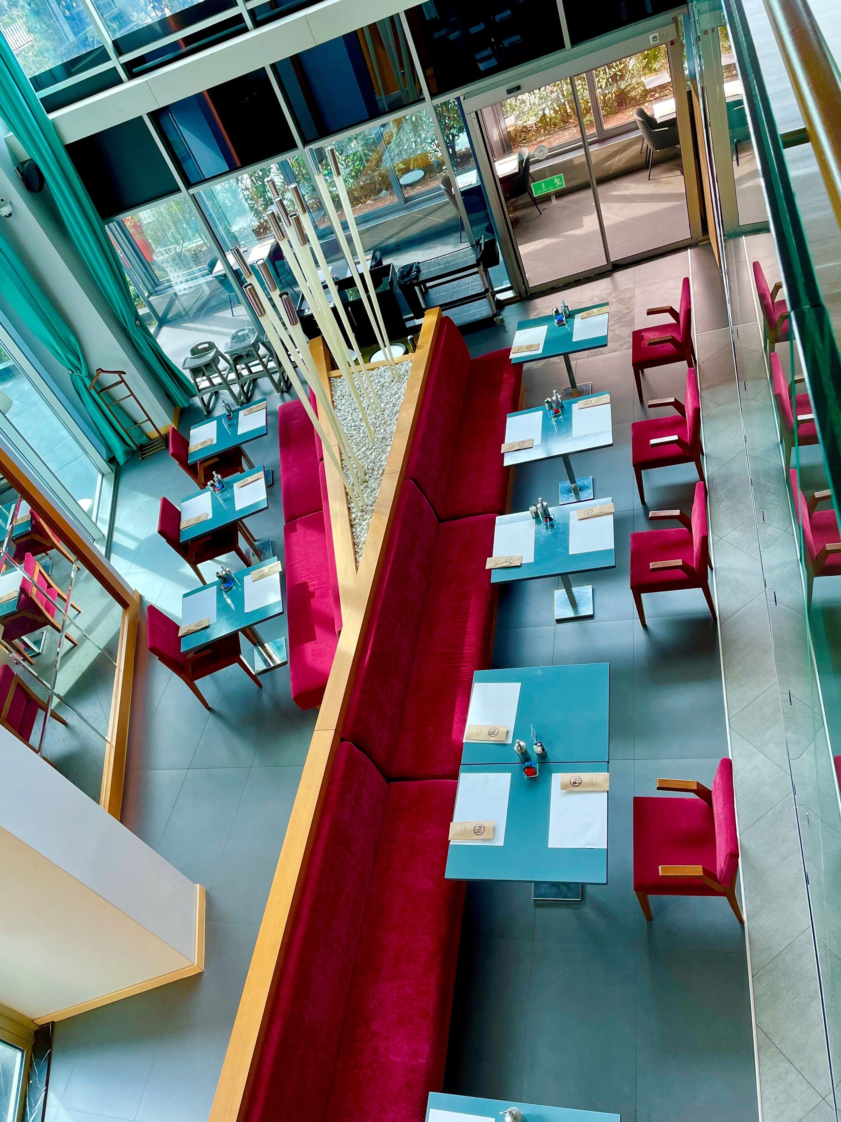 Un área de estar interior moderna con bancos acolchados de color rojo y mesas azules, que cuenta con separadores de vidrio y luz natural que entra por amplias ventanas.