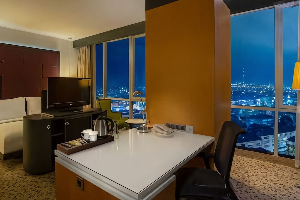 A hotel room with a city view, a desk with a telephone, a television, and a seating area with a chair and a small table.