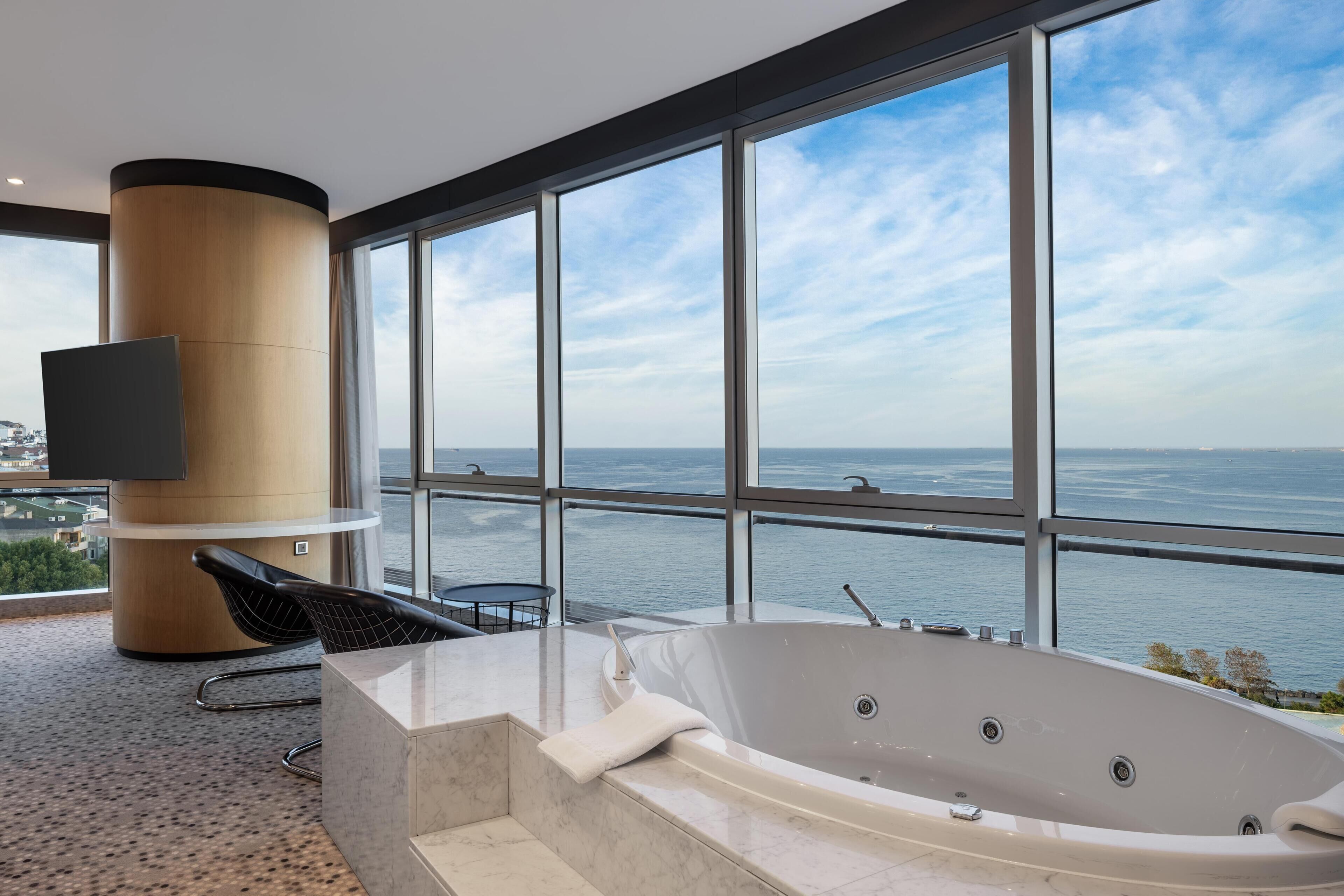 A modern bathroom with a large bathtub, a flat-screen TV, and a view of the sea.