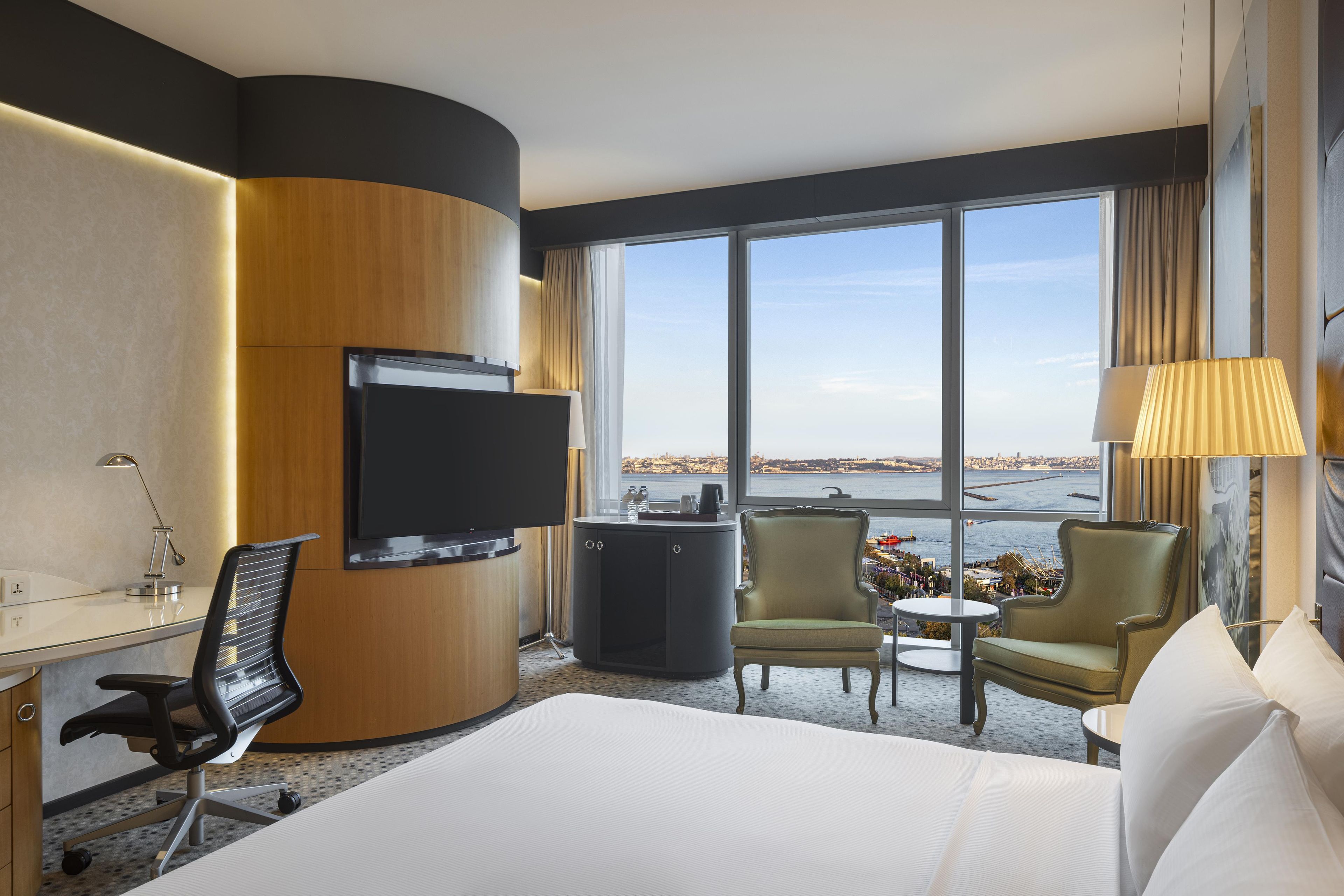Habitación de hotel con cama, escritorio, televisor, silla, mesita y vista a la ciudad a través de amplios ventanales.