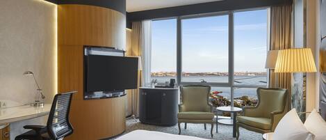 Habitación de hotel con cama, escritorio, televisor, silla, mesita y vista a la ciudad a través de amplios ventanales.