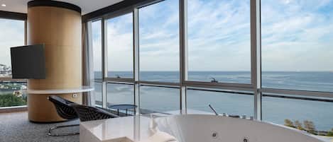 A modern bathroom with a large bathtub, a flat-screen TV, and a view of the sea.