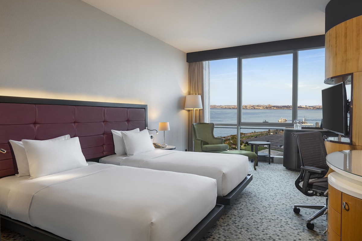 Habitación de hotel con dos camas, un escritorio, una silla, un televisor y vista al mar.