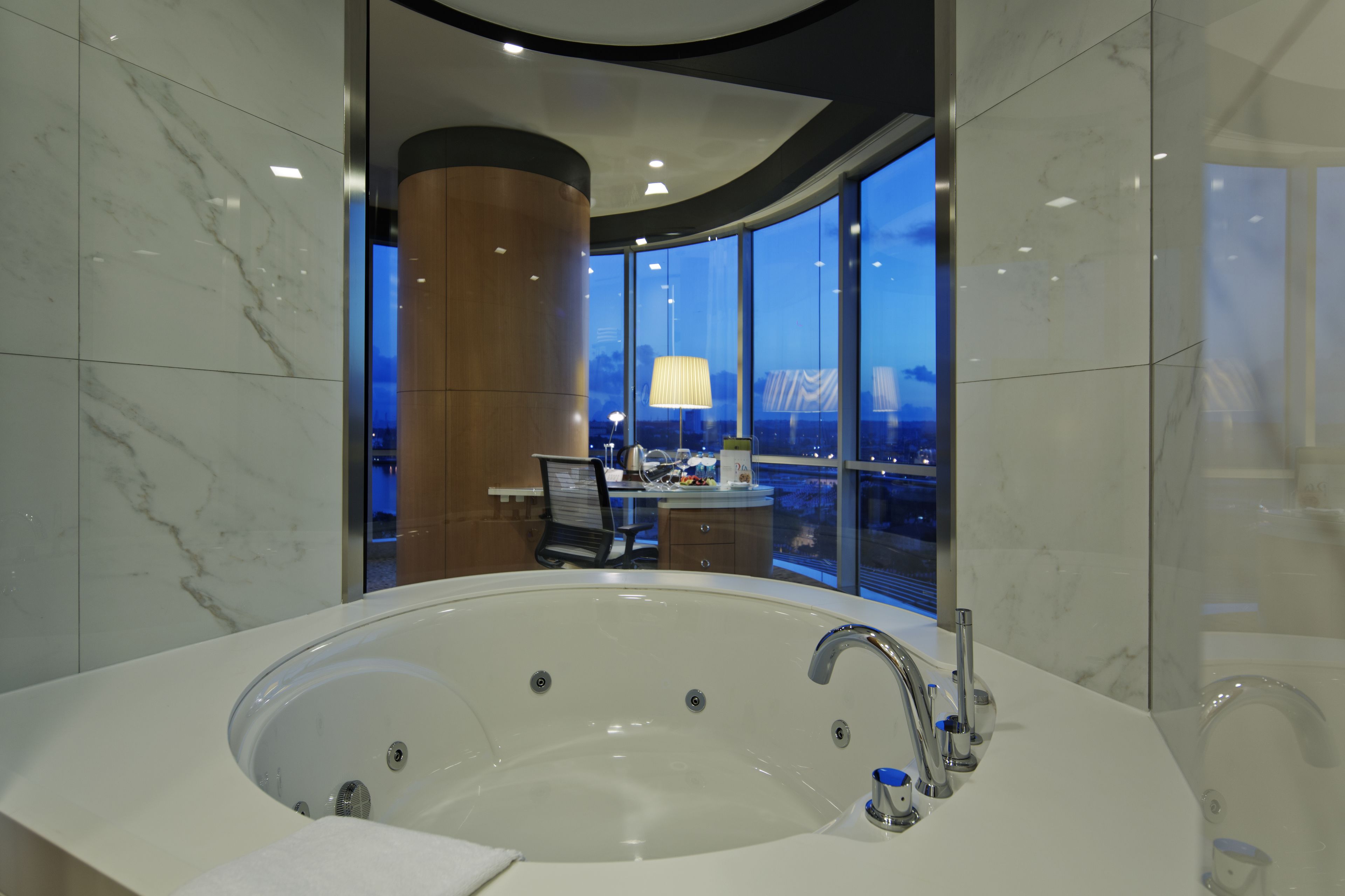 A modern bathroom with a large bathtub, a desk with a chair, and a view of the cityscape at dusk.