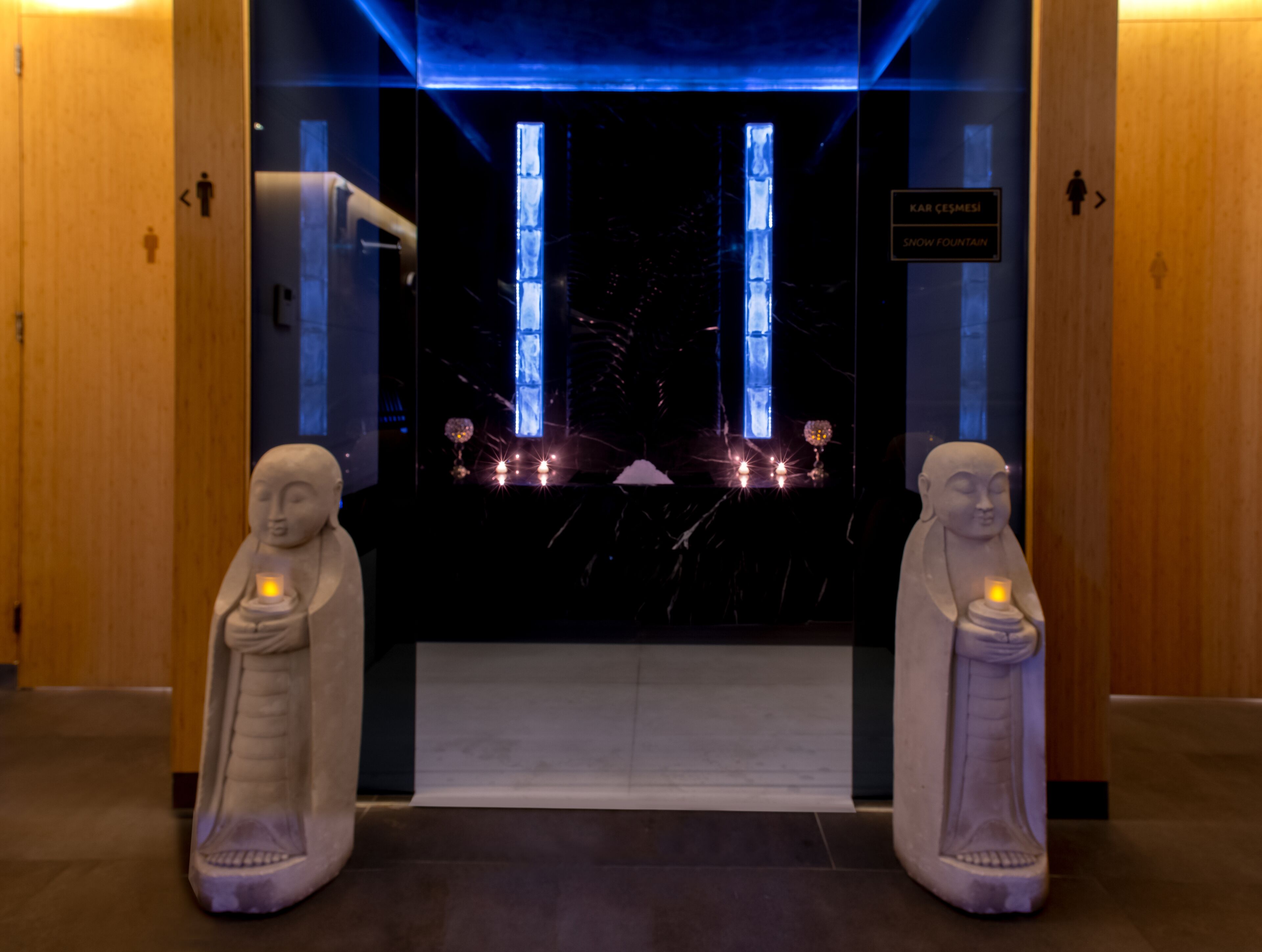 A hallway with two statues of Buddha, a blue-lit wall, and a sign indicating the direction to the Snow Fountain.