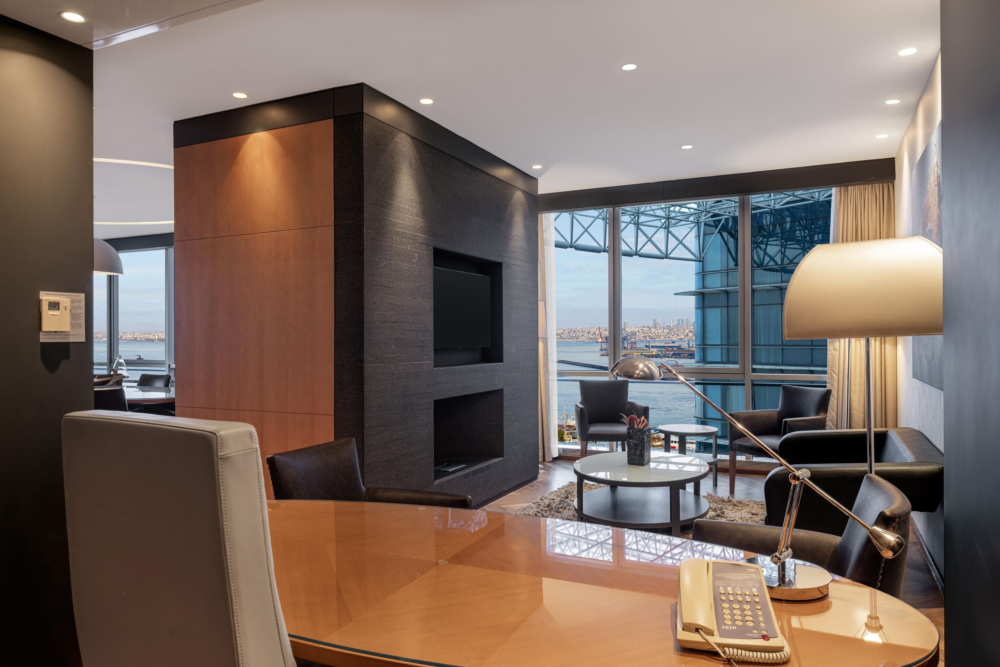 A modern hotel room with a large window, a flat-screen TV, a desk with a telephone, and a seating area with a lamp.