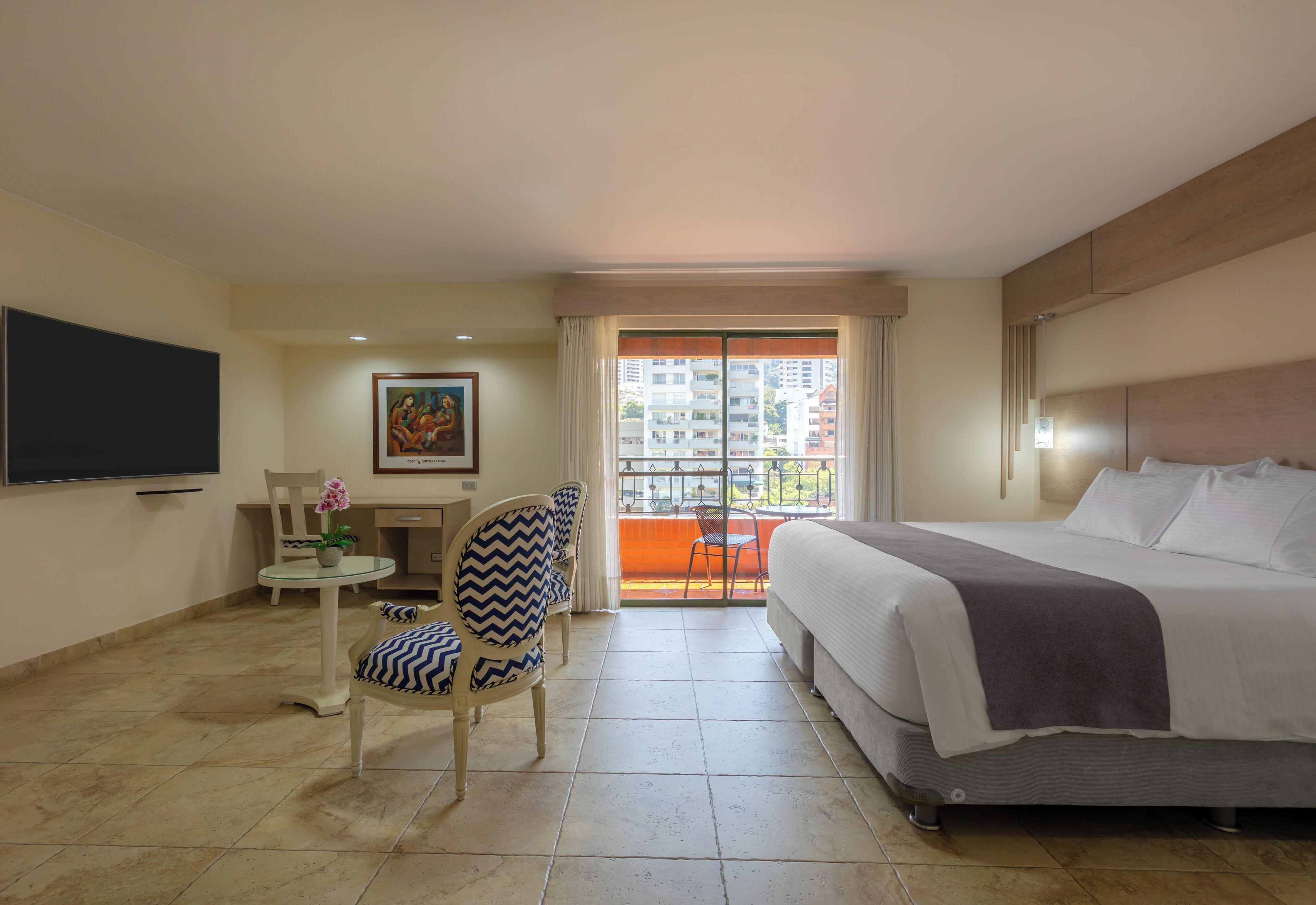 A hotel room with a bed, a chair, a small table with a vase of flowers, a painting on the wall, and a balcony with a view of buildings.