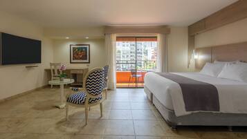 A hotel room with a bed, a chair, a small table with a vase of flowers, a painting on the wall, and a balcony with a view of buildings.