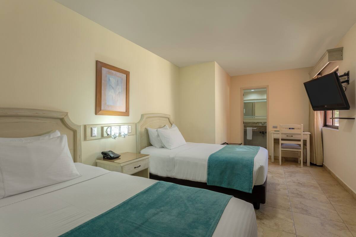 Habitación de hotel con dos camas, un escritorio, una silla, un televisor y un baño visible a través de una puerta abierta.