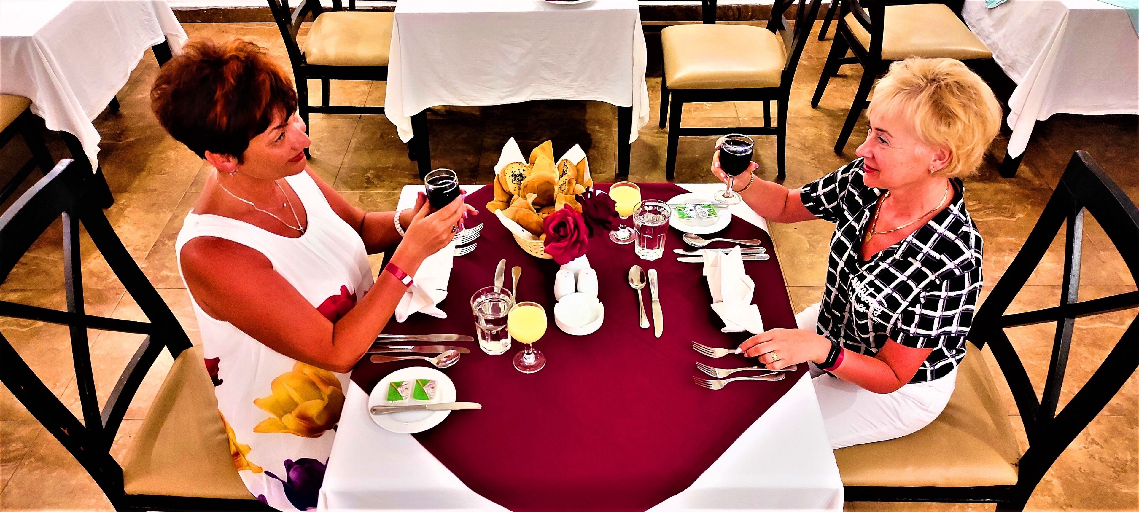 Two people seated at a table with a red tablecloth, white tablecloths in the background, and a floral pattern on the chair's cushion.