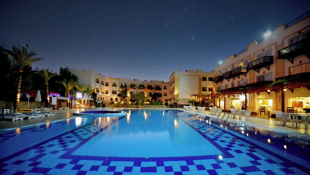 Una piscina con un diseño geométrico de azulejos, rodeada de palmeras y edificios con balcones.