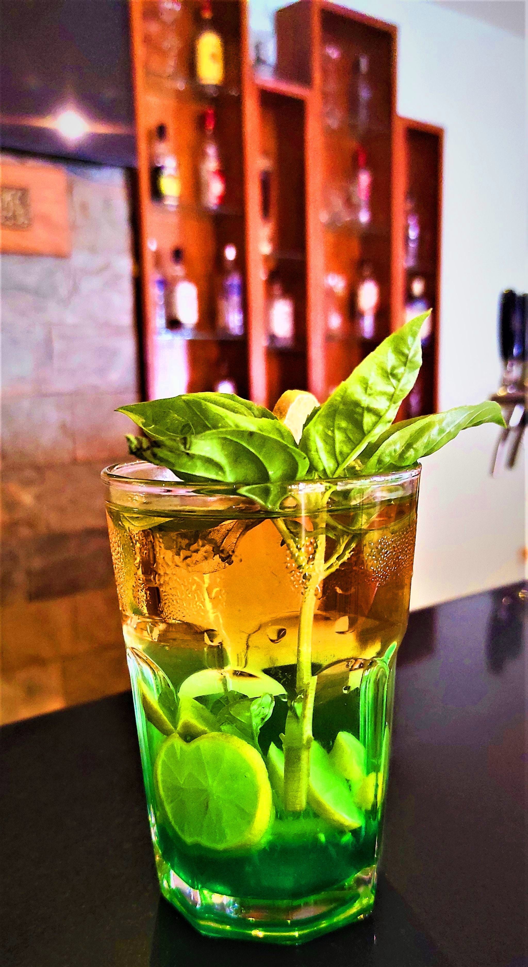 Un vaso de bebida verde con rodajas de lima y una hoja de albahaca, sobre una barra con un fondo borroso de estantes llenos de botellas.