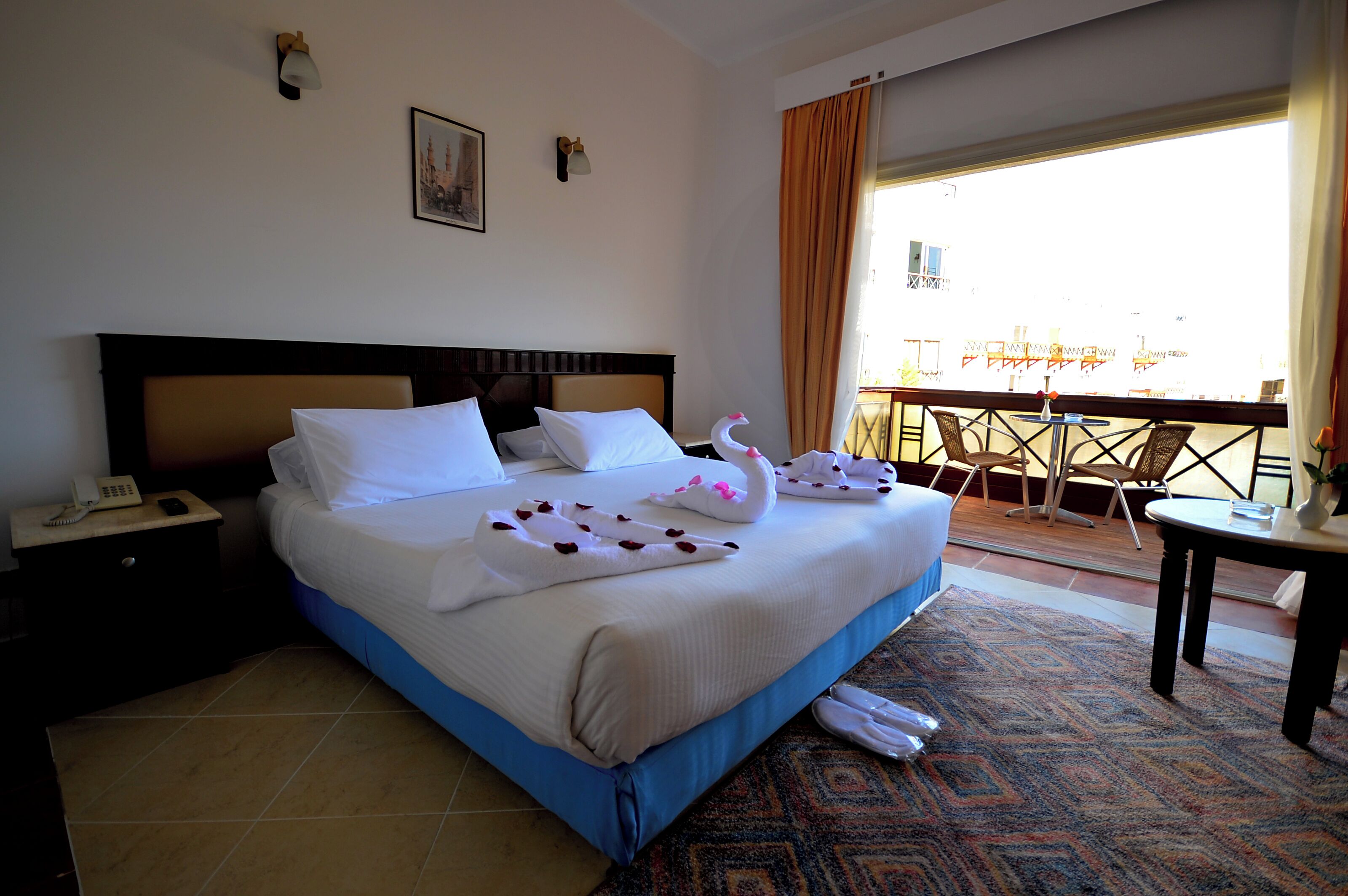 A hotel room with a large bed, a nightstand with a phone, a balcony with chairs and a table, and a patterned rug.